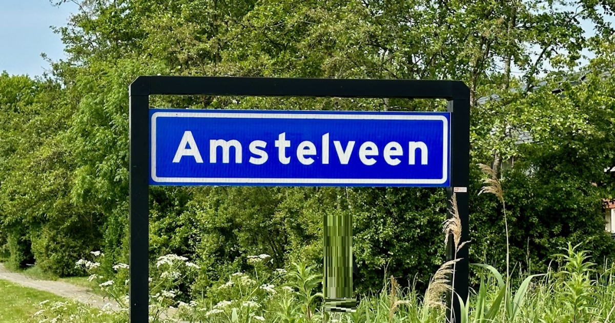 Blue town sign reading Amstelveen in white letters, mounted on a black frame with green trees behind it